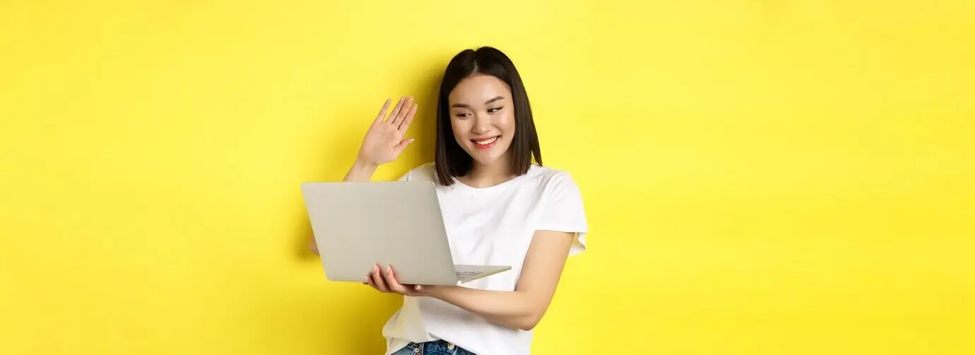 Vrolijke Aziatische vrouw die op een laptop videobelt, naar de computercamera zwaait en hallo zegt, terwijl ze staat.