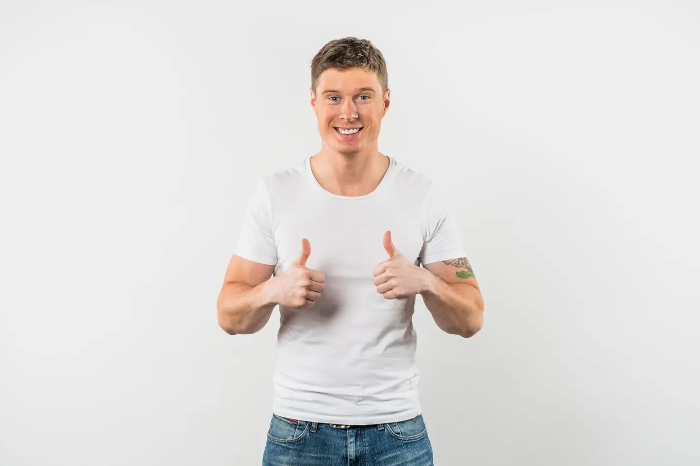 Lachende jonge man die met beide handen een duim omhoog steekt tegen een witte achtergrond.